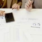 Dua profesional menganalisis dokumen keuangan dengan kalkulator untuk keperluan manajemen akuntansi